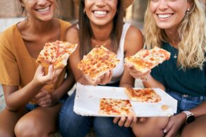 Close up of pizza. Three beautiful women sitting on the stairs of the city eating street food